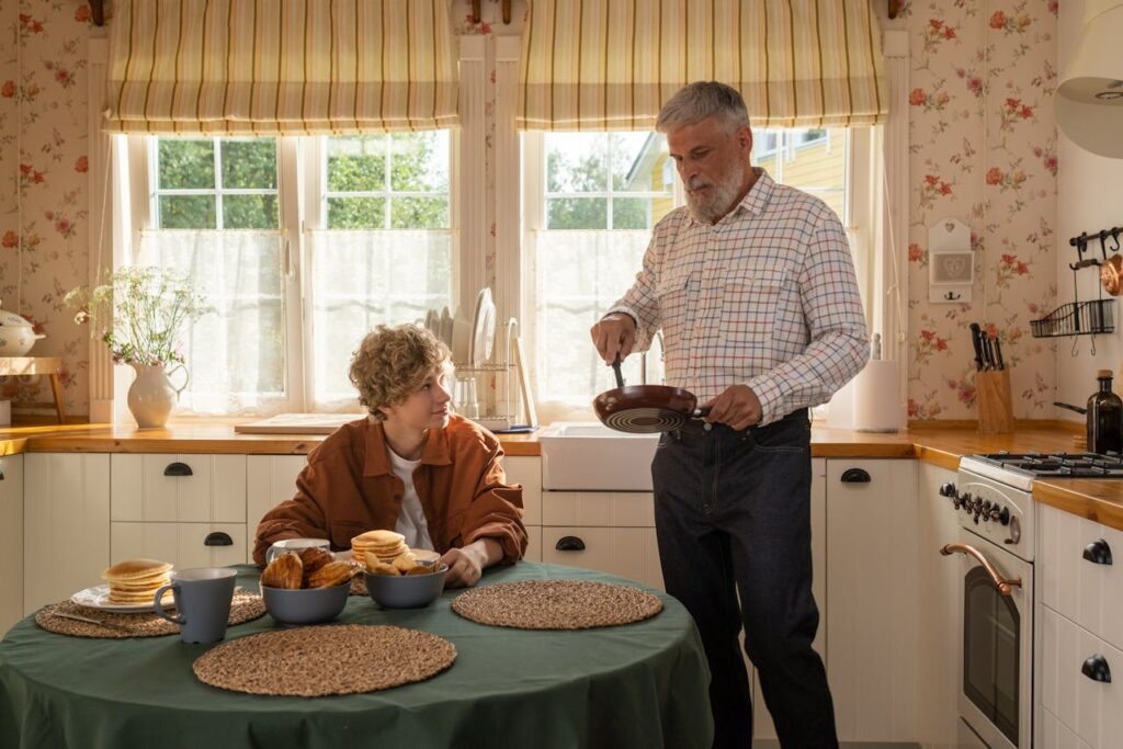 Bild mit einer weißen Küche im Landhausstil und einem Mann, der Pfannkuchen macht und einem Jungen, der am Küchentisch sitzt